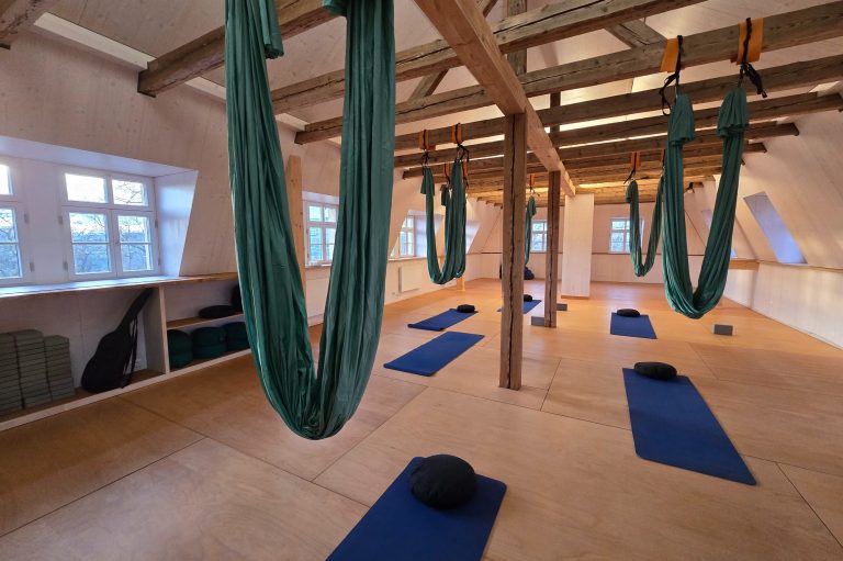 Yoga-Raum mit sichtbaren Balken und hängenden Yoga-Tüchern, Matten und Kissen auf dem Boden.